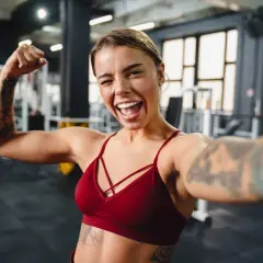 Uśmiechnięta kobieta z tatuażami pokazuje biceps po ćwiczeniach na siłowni.