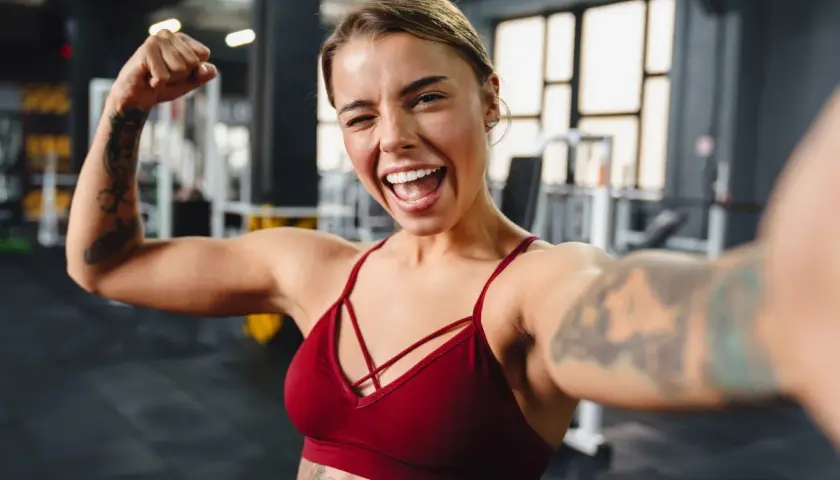 Uśmiechnięta kobieta z tatuażami pokazuje biceps po ćwiczeniach na siłowni.