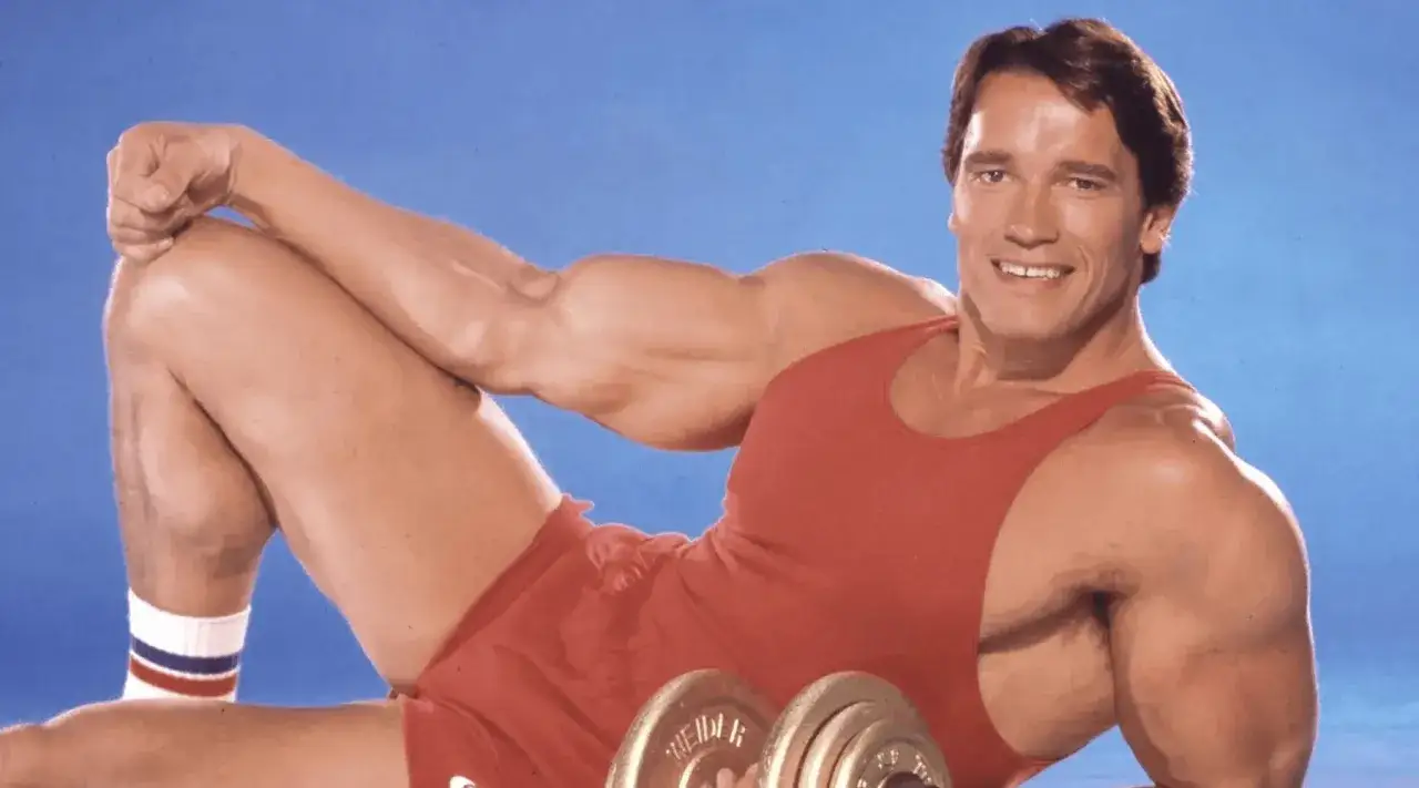Arnold Schwarzenegger w czerwonym trykocie i szortach, z hantlami, prezentuje swoją muskulaturę. Jego słynny
