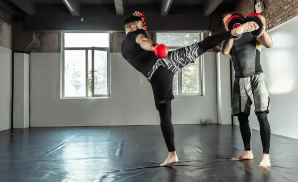 Trener łapie kopnięcie low kick. Dwóch mężczyzn w rękawicach treningowych ćwiczy sztuki walki na sali.